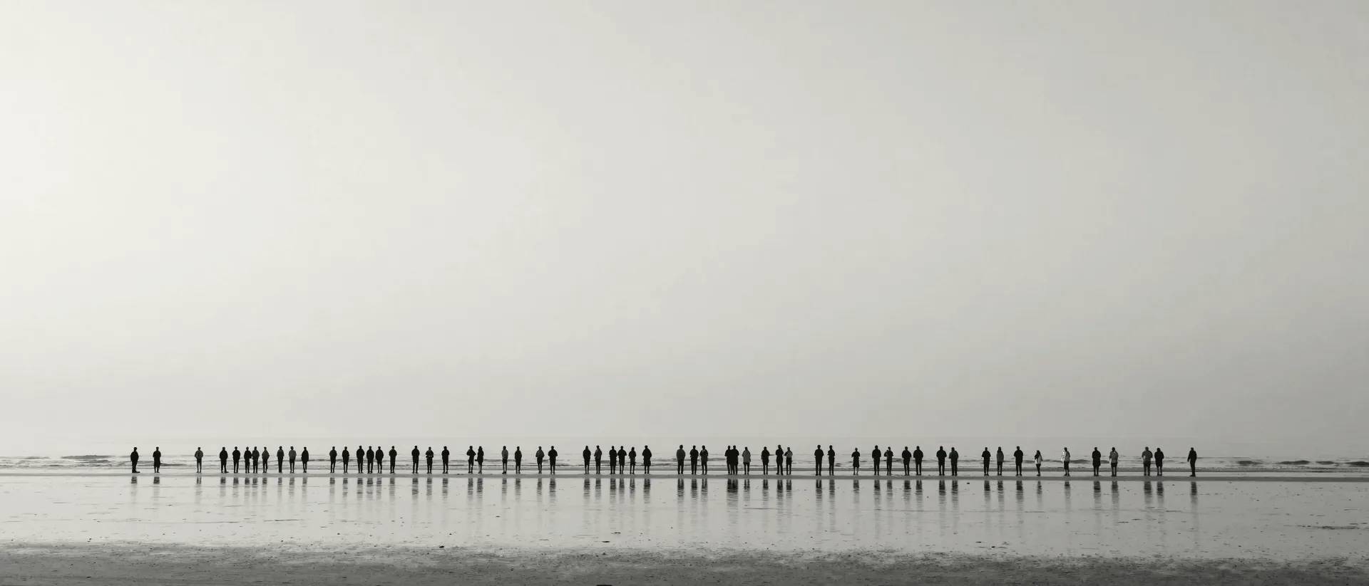 A long line of people standing far apart along a flat, empty shoreline under a wide sky.